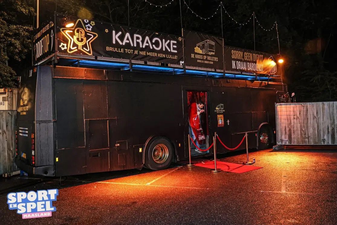 De Kroegbus in de avond met karaokebanners op het dak, rode loper en warme verlichting.