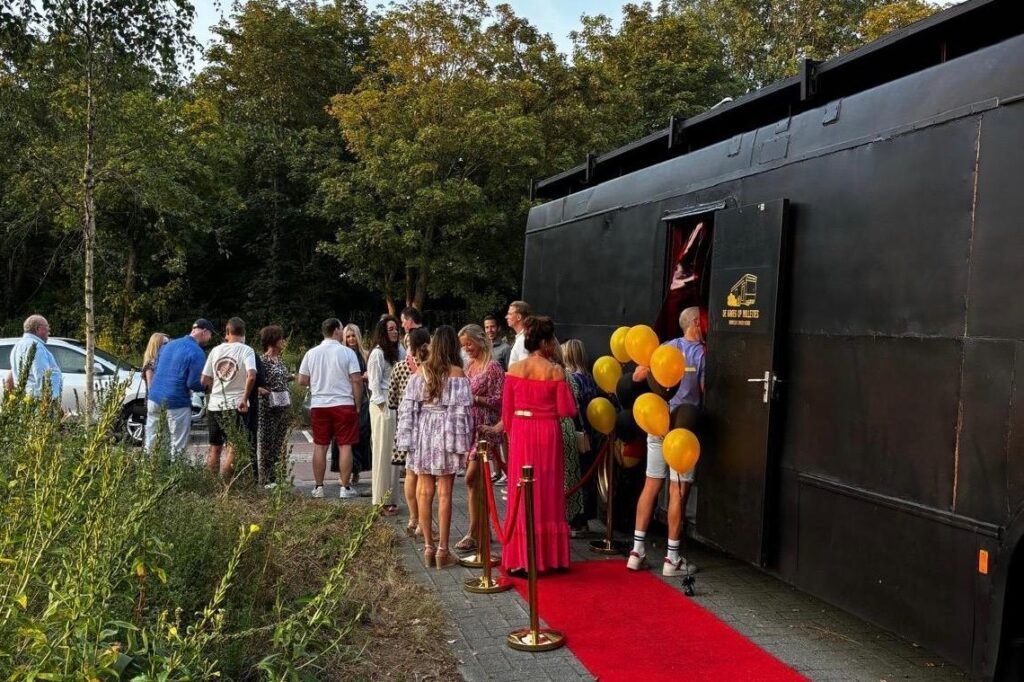 Gasten op de rode loper bij de bus met gouden en zwarte ballonnen.